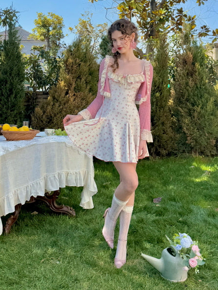 A model wearing a vintage-inspired floral dress with a soft pink bolero featuring delicate lace trim, standing outdoors beside a table adorned with a lace tablecloth and a basket of fresh lemons.