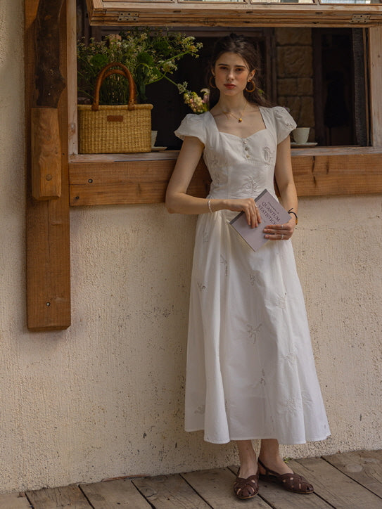 Woman leaning against a rustic wooden window frame, holding a book while wearing a white, floral-embroidered long dress with a soft, romantic silhouette.