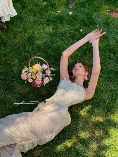 Woman wearing cream ruched satin midi skirt and floral corset top, lying on grass with arms stretched and a basket of flowers beside – romantic fairytale garden fashion