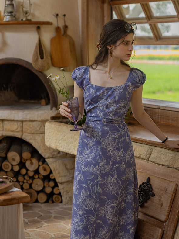 Young woman in a blue floral dress holding a glass with wildflowers, standing by a stone kitchen counter with a firewood oven in a cozy cottage setting.