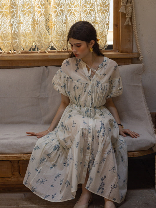 Woman sitting on a wooden sofa in a cream floral print dress with blue botanical patterns, surrounded by lace curtains. The cottagecore-inspired setting evokes a serene, vintage atmosphere.