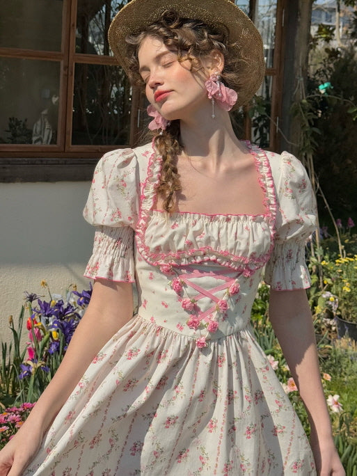 A young woman in a pink floral cotton dress poses under sunlight, wearing a straw bonnet and large flower earrings. The vintage-inspired dress features a square neckline, puffy sleeves, and a ribbon-laced bodice with frill details, perfectly matching the romantic fairytale aesthetic of a spring garden.
