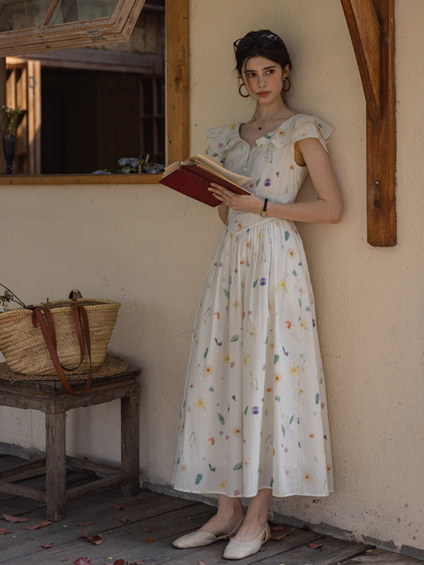 Woman leaning against a rustic wall, holding a book while wearing a cottagecore-inspired floral dress with ruffled sleeves, a woven basket resting nearby.