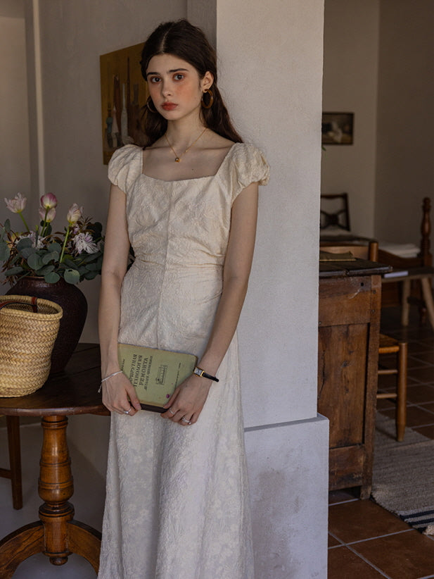 Young woman in a cream-colored textured midi dress with puff sleeves and a square neckline, holding a vintage book while standing against a wall. A woven basket and a vase of flowers create a cottagecore-inspired atmosphere.