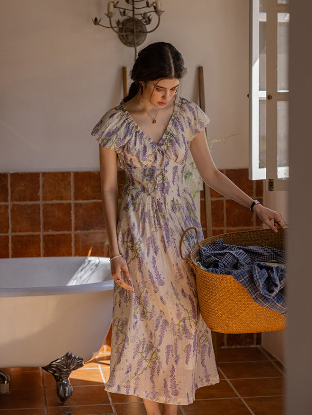 Woman carrying a woven basket of linens while wearing a lavender floral cottagecore dress, set in a vintage-style bathroom with warm terracotta tiles and a clawfoot bathtub.