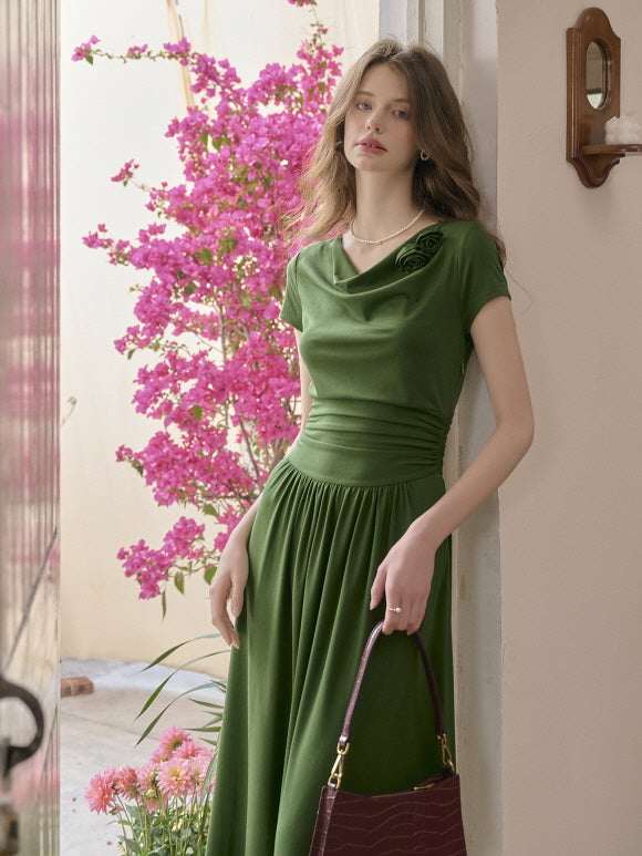 Model wearing a soft green dress with a gathered waist and a flower brooch detail, leaning against a wall near blooming pink flowers. Graceful countryside elegance with a dreamy atmosphere.