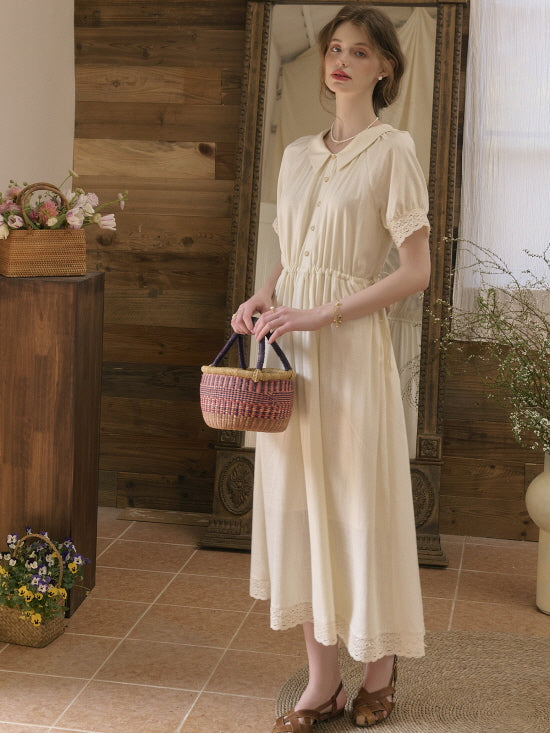 Model standing indoors holding a woven basket, wearing a cream vintage dress with lace sleeves and button-down front.