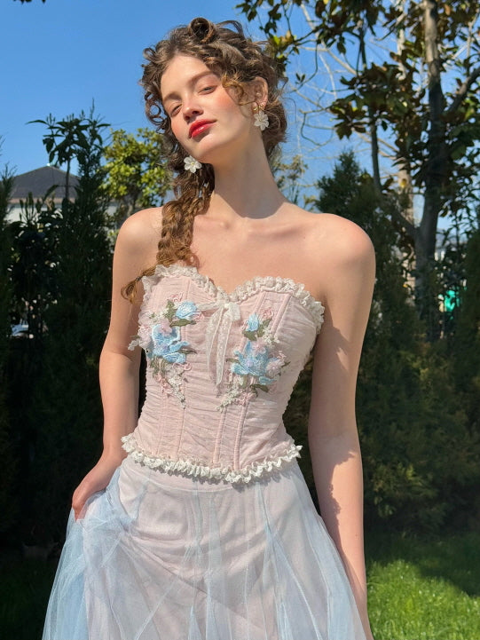 Front view of pastel pink floral corset dress with blue embroidery and tulle skirt, worn by a woman in a fairytale garden – elegant vintage-inspired outfit