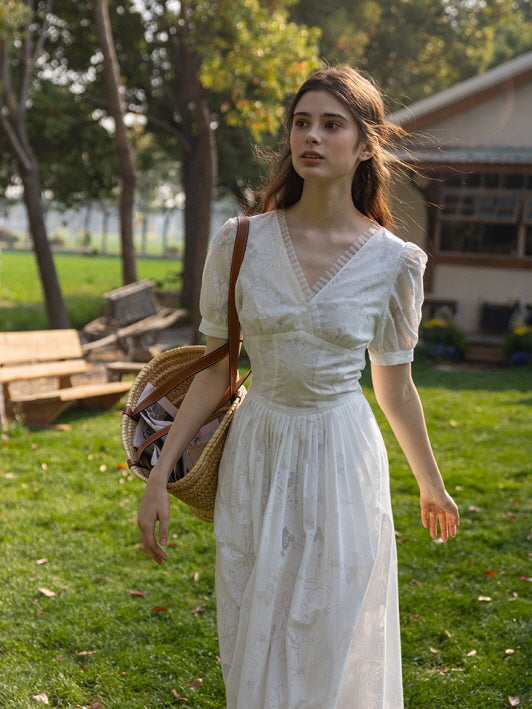 Woman in a white floral embossed midi dress walking through a lush green lawn, carrying a woven tote bag, with a wooden shed and flower beds in the background.
