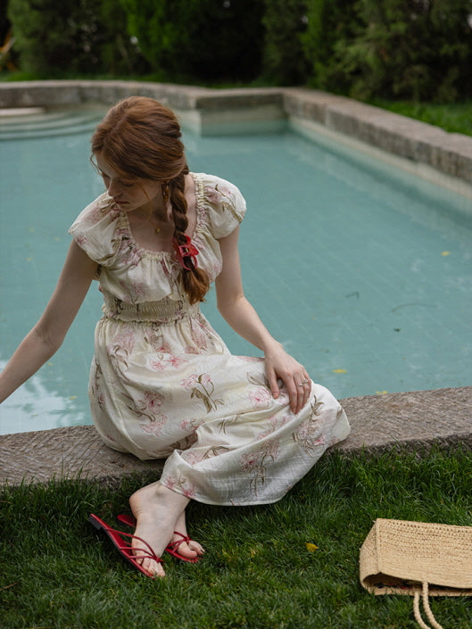 A woman sitting near a pool, gently touching the grass while wearing a floral cottagecore dress with cap sleeves and red sandals. Vintage-inspired, fairytale look.