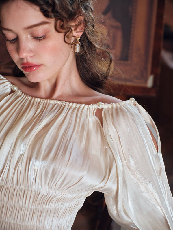 A woman with soft curls gazes downward while wearing a pearl-detailed cream blouse with delicate pleats, evoking a timeless, romantic vintage mood in a painterly setting.