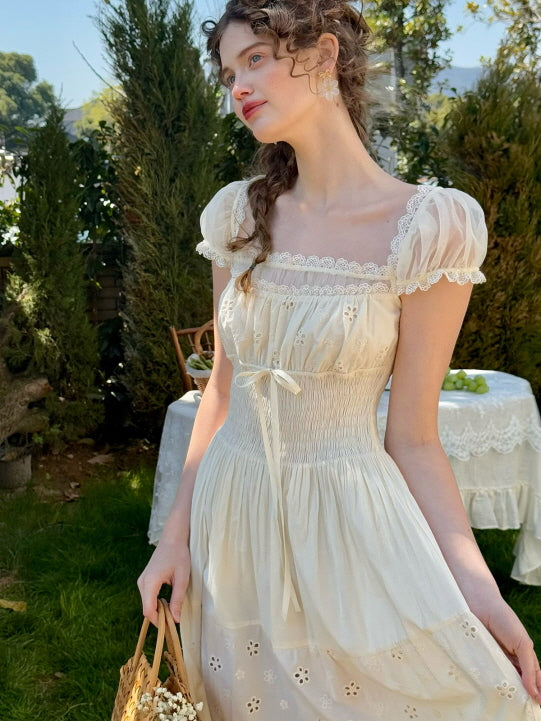 A woman standing in a garden wearing a romantic ivory dress with puff sleeves, lace trim, and floral embroidery, holding a woven basket in a soft countryside setting.