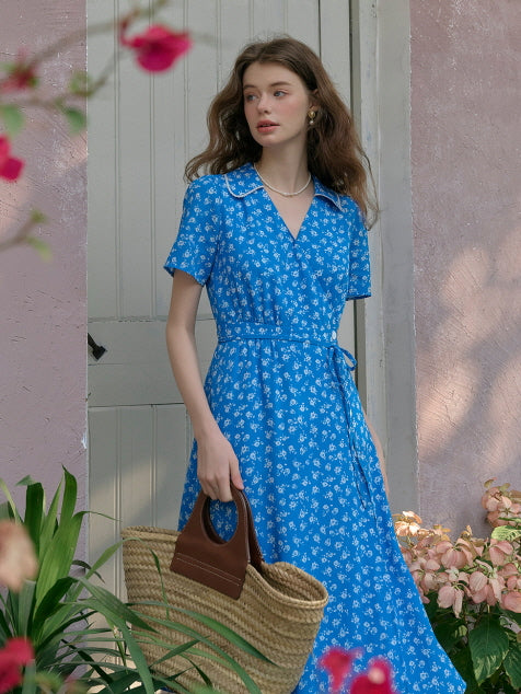 Model wearing a blue floral belted dress, holding a straw bag, standing in front of a rustic door with blooming flowers. cottagecore garden outfit with fairytale-inspired charm.