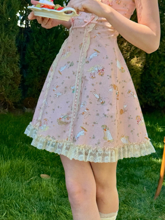 Close-up of a pink vintage-style dress adorned with whimsical fairytale illustrations and delicate lace trim, as a young woman holds a plate of strawberry pastries.