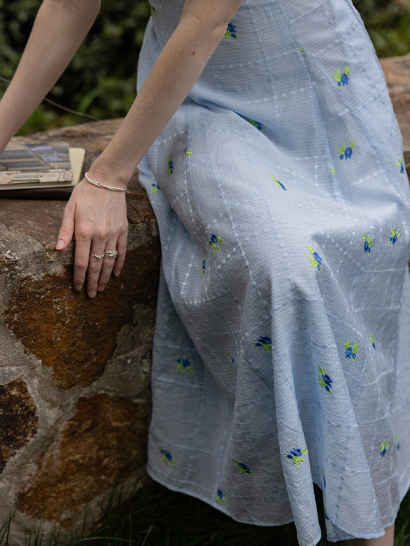 Close-up of a blue floral midi dress with delicate embroidered flower patterns and a soft, flowy texture, paired with silver rings and a rustic stone backdrop.