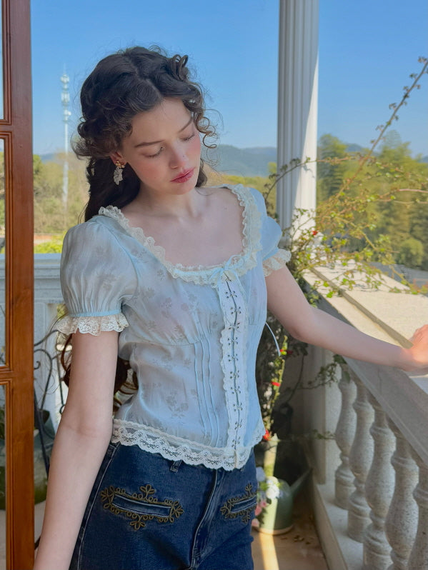 A model leans against a balcony rail, wearing a pastel blue floral corset top with lace detailing and puff sleeves, paired with dark denim jeans featuring floral embroidery, capturing a soft, vintage-inspired aesthetic.