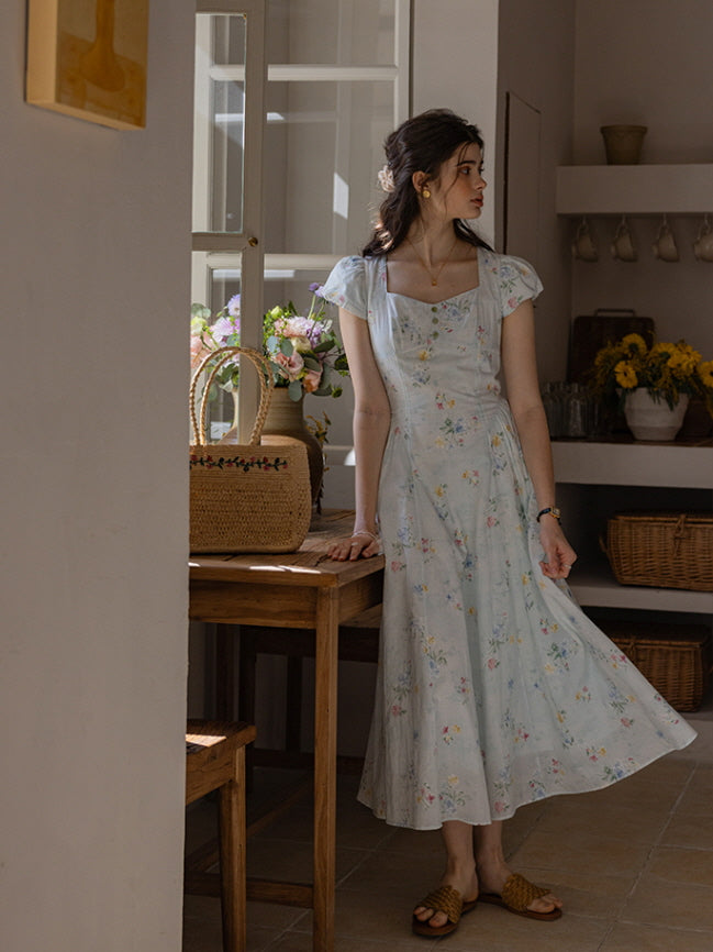 Romantic floral dress with a flowing silhouette and delicate button-down bodice, paired with straw sandals and a woven floral-embroidered bag in a rustic, cottagecore kitchen setting.