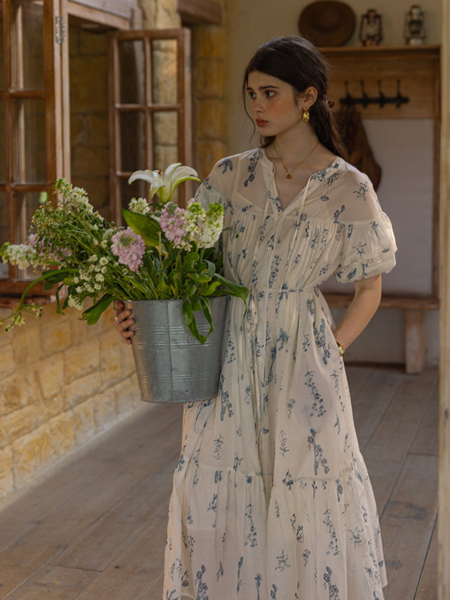 Young woman in a cream floral print dress with tiered skirt and puff sleeves, holding a large metal bucket filled with fresh flowers while standing on a rustic wooden porch. The scene exudes a romantic, countryside aesthetic.