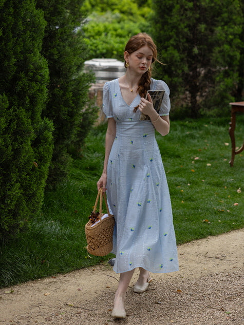 Woman in a blue floral midi dress walking through a garden path, holding a woven basket and a book, exuding a serene, cottagecore summer aesthetic.