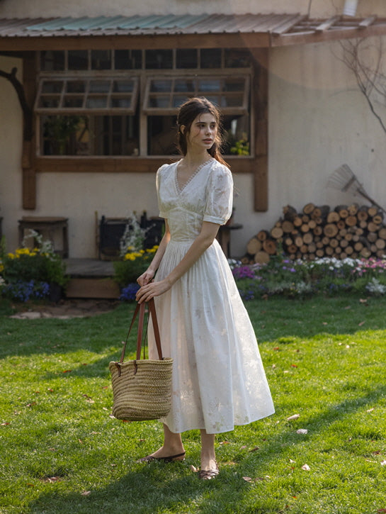 Side view of a woman in a white floral embossed midi dress walking with a serene expression, holding a woven tote bag under her arm in a garden setting.