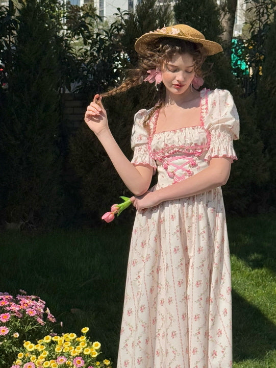 A model stands in a blooming garden holding a tulip, wearing a puff-sleeved vintage-style dress with delicate pink flower patterns, corset-like ribbon detailing on the bodice, and soft shirring. The look expresses the dreamy charm of cottagecore and Romantic Fairytale fashion.