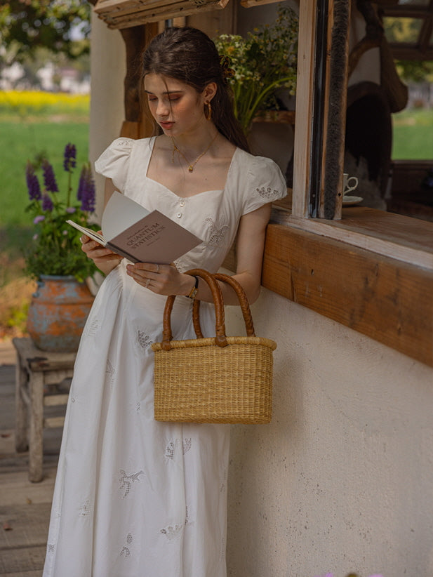 Woman standing by a rustic wooden window wearing a white embroidered cotton dress, holding a book and a woven basket bag, evoking a vintage-inspired countryside aesthetic.