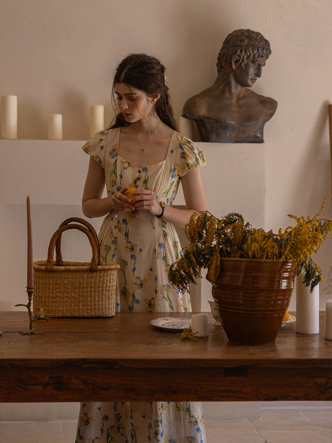 The model stands gracefully at a wooden dining table, holding a piece of fruit as she gazes downward. Her cream floral dress with blue and yellow flower prints flows elegantly, complemented by a woven basket bag and a classical bust sculpture in the background, creating a dreamy, old-world atmosphere.