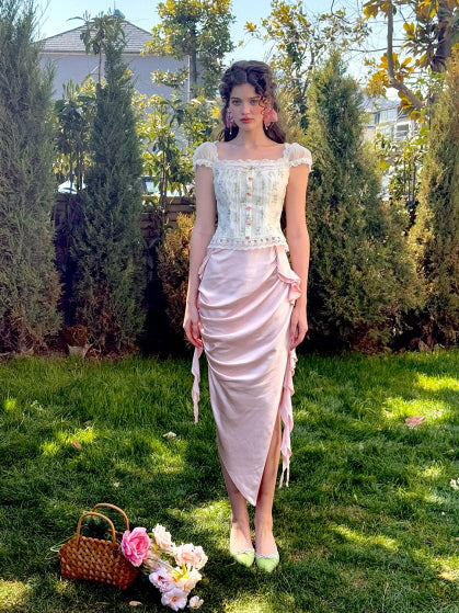 A model stands in a garden wearing a delicate floral corset top with lace detailing and off-shoulder puff sleeves, paired with a satin ruched pink skirt adorned with ribbon ties, creating a romantic, fairytale-inspired look.