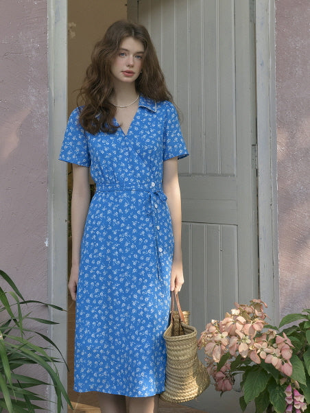 Young woman standing calmly in a vintage-style blue floral dress with waist tie, holding a straw basket bag at a pastel pink doorway. Elegant princesscore outfit with soft garden charm.