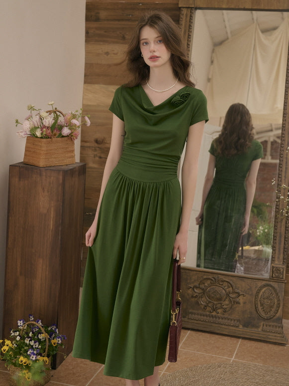 Woman standing gracefully in a forest green draped-neck dress with a cinched waist, holding a small handbag. Romantic indoor setting with a vintage mirror and floral accents.