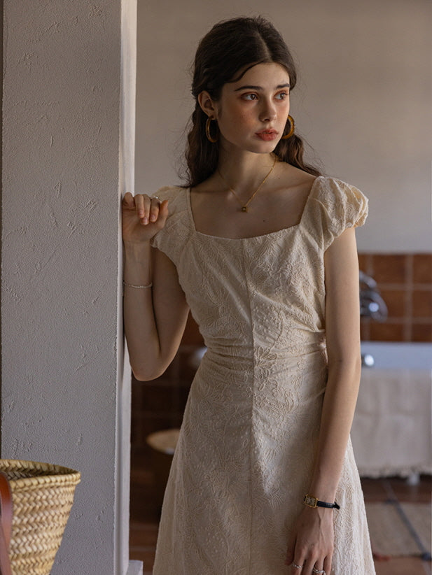 Woman wearing a cream-colored textured midi dress with a square neckline and puff sleeves, leaning against a wall while gazing thoughtfully to the side in a softly lit, vintage-inspired room.