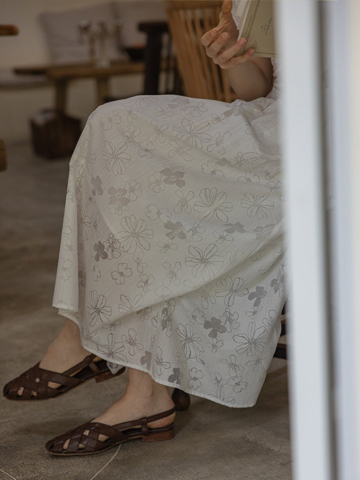 Detailed view of the floral-patterned skirt of a white cotton dress, with intricate flower embroidery and a flowy silhouette, paired with brown woven sandals.