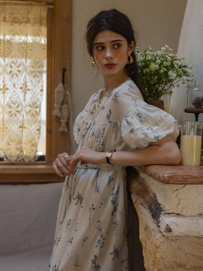 Young woman in a cream floral print dress with delicate blue floral patterns, leaning against a rustic stone mantel while gazing thoughtfully to the side. The soft, cottagecore setting includes lace curtains and wildflowers.