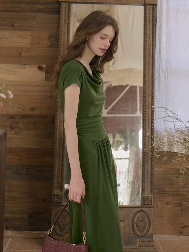 Side view of a woman wearing a forest green midi dress with a draped neckline and gathered waist, holding a burgundy handbag. Soft vintage mood reflected through natural lighting and rustic decor.