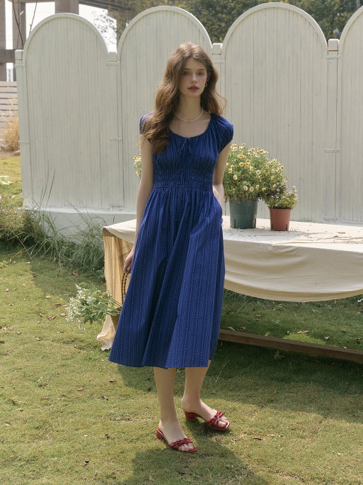 Model wearing a shirred navy dress with a flowy midi silhouette, standing on a grassy lawn with floral arrangements and vintage garden décor. Elegant countryside charm with romantic details.
