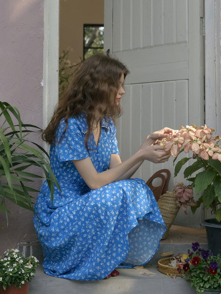 Woman in a blue floral dress gently touching pink flowers while sitting by a rustic doorway. Romantic and serene cottagecore scene with natural light and garden details.