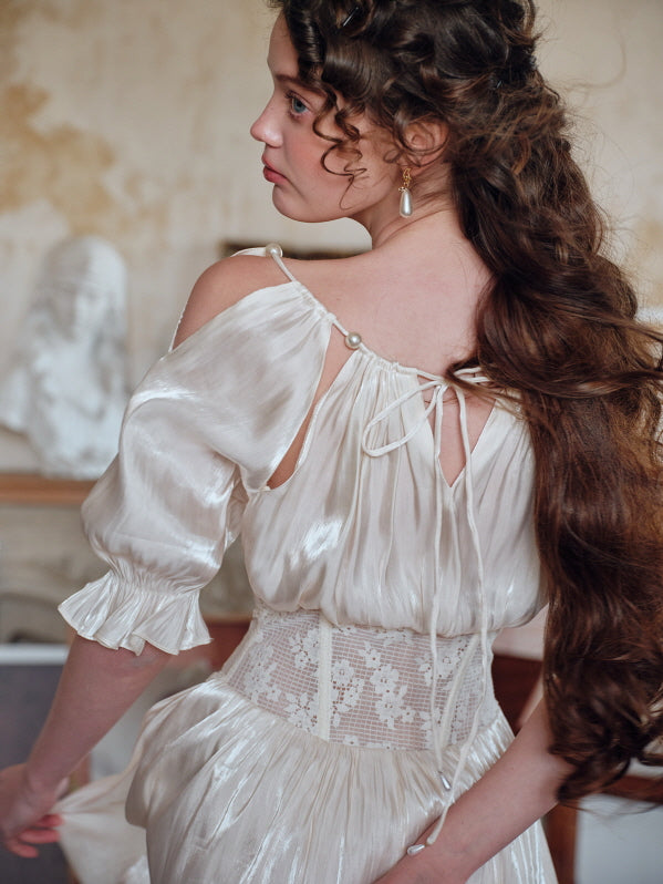 Back view of a woman wearing a pearl-accented ivory dress with open shoulder details and a lace corset, standing in a vintage studio setting with long curled hair cascading down her back.