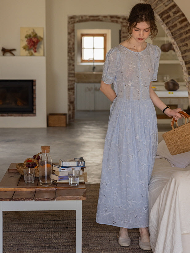 A woman standing near a rustic wooden table, holding a woven basket while wearing a blue eyelet lace dress with puff sleeves. Cozy cottagecore setting.