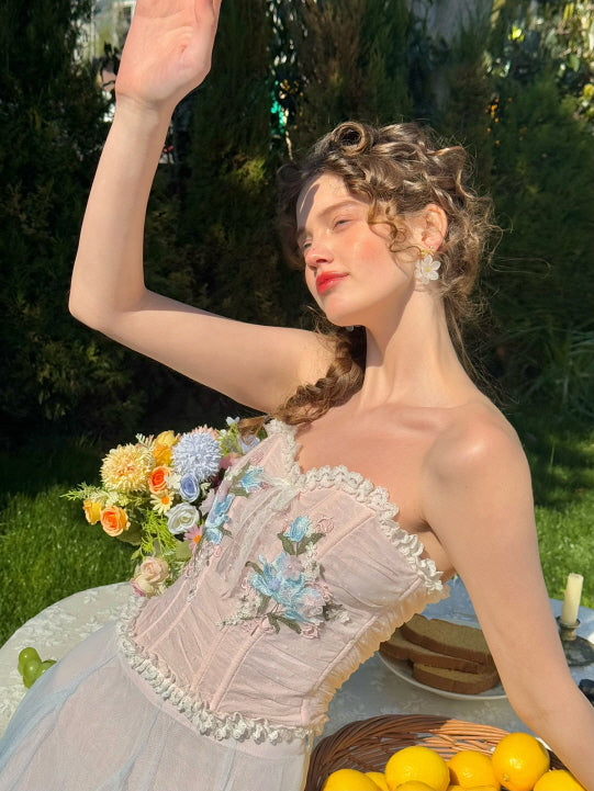 Romantic pastel corset dress with embroidered blue flowers, worn in a garden picnic setting with lemons and fresh floral bouquet – fairytale spring fashion for women
