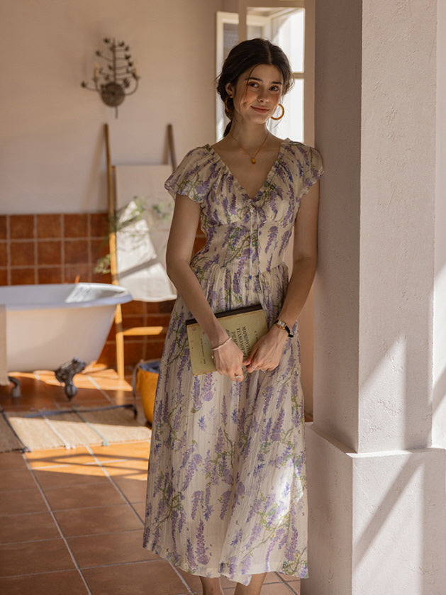 Woman in a lavender floral dress leaning against a white pillar, holding a book in a serene cottagecore-inspired bathroom with warm terracotta tiles and a vintage clawfoot tub.
