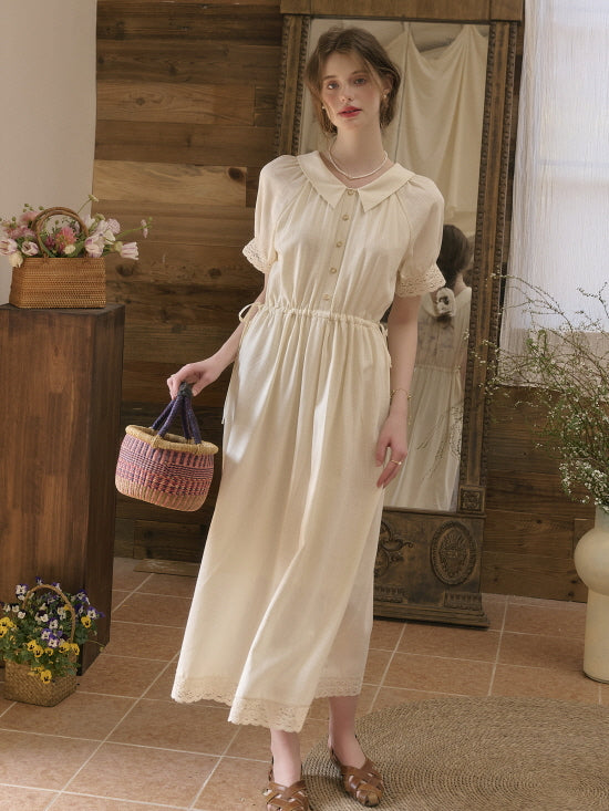 A woman wearing a vintage cream dress with lace trim and a peter pan collar, holding a straw basket, standing in a cozy rustic interior.