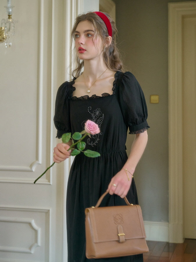 Retro-inspired vintage black dress with floral embroidery, puff sleeves, and lace trim, styled with a red headband and brown handbag, model walking elegantly in a classic hallway.