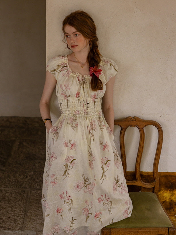 A woman standing near a vintage wooden chair, dressed in a cottagecore floral dress with a delicate pink and green pattern, exuding a romantic, fairytale look.