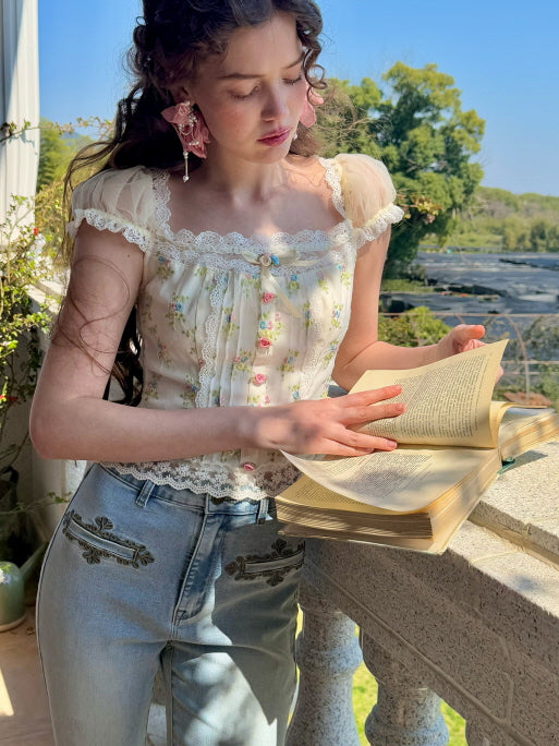 A model leans against a balcony while reading a book, dressed in a floral corset top with lace trim and sheer puff sleeves, styled with embroidered denim jeans, evoking a soft, vintage aesthetic.