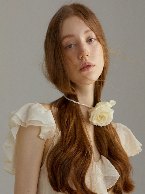 Close-up portrait of a model wearing an ivory ruffle dress with sheer shoulder details, accented by a delicate rose choker and soft natural makeup for a romantic, ethereal mood.