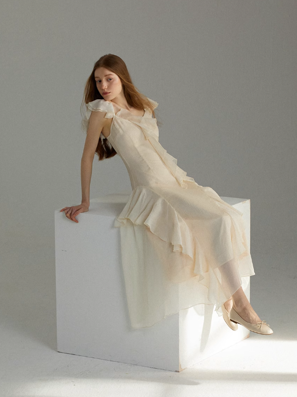 Full-length ivory chiffon dress styled on a seated pose, showcasing cascading ruffle details, a softly fitted bodice, and a dreamy fairytale-inspired look in a clean studio environment.