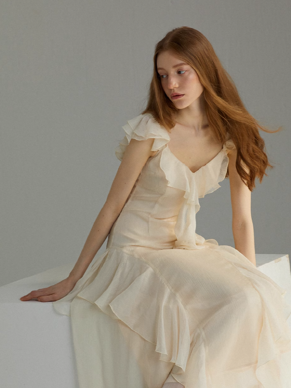 A romantic ivory chiffon dress with soft ruffle sleeves and layered flowing skirt, worn by a model seated on a minimalist studio block, showcasing an airy silhouette and delicate movement.