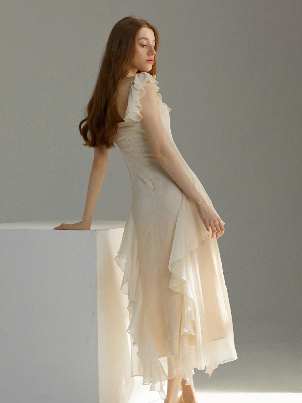 Soft ivory chiffon dress shown in a side profile, featuring layered ruffle sleeves and an asymmetrical flowing hem, styled with a gentle lean against a white cube in a minimalist studio setting.