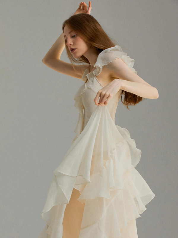 A flowing ivory chiffon dress captured in motion, featuring layered ruffle skirt details and soft flutter sleeves, styled in a minimalist studio setting with a graceful, airy silhouette.