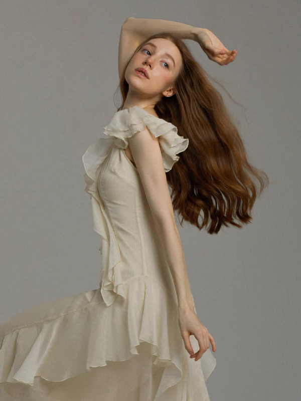 Close-up of an ivory ruffle chiffon dress with flutter sleeves and soft draping, emphasizing graceful movement, flowing hair, and a light, ethereal silhouette against a neutral backdrop.